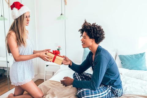 A joyful scene of a couple exchanging a wrapped gift in a cozy bedroom. The woman wears a Santa