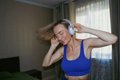A woman is joyfully dancing at home, wearing white headphones. Her eyes are closed as she moves to