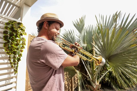 A talented one-armed man wearing a hat plays a trumpet outdoors, surrounded by greenery and