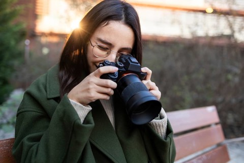 A woman wearing a green jacket is sitting on a park bench, smiling as she takes photos with a
