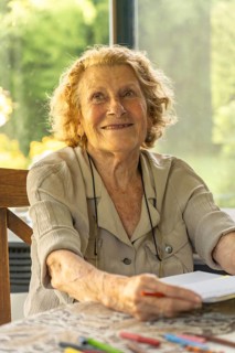 Happy elderly woman sitting at a table near a window, drawing on a notepad with colored pencils,