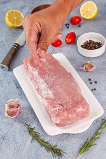 A hand seasons a raw pork loin with salt on a plate. Surrounding ingredients include garlic,
