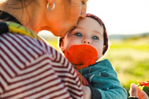 A mother tenderly kisses her daughter on the head while the child holds a vibrant poppy in a sunny