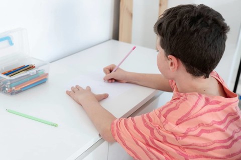 A young boy focused on drawing at a white table, surrounded by colorful pencils and school supplies