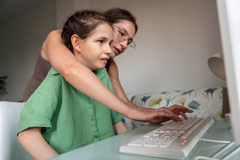 A sister shares a moment as she helps a girl with a tracheostomy use a computer. This scene