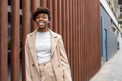 A stylish black woman smiles confidently while leaning against a wooden fence in a modern urban