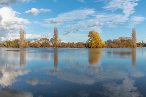 The river flows flat and reflects clouds and mountains under a clear sky. Autumn trees draw the