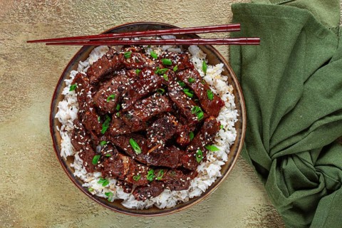 Crispy beef, with green onions and sesame seeds, on a rice pad, in a bowl with chopsticks, on a