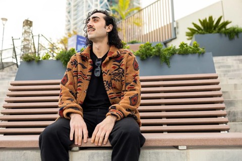 A man with curly hair sits on a bench in an urban setting, wearing a patterned jacket and black