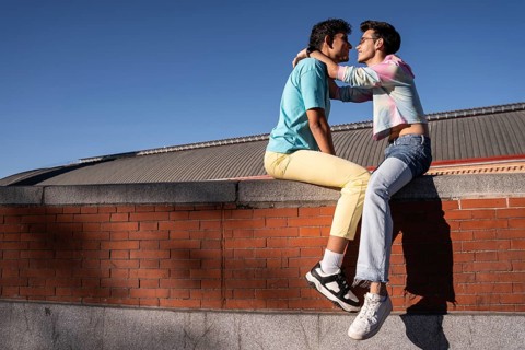 A joyful moment shared by a gay couple, sitting on a brick wall under a clear blue sky. Their