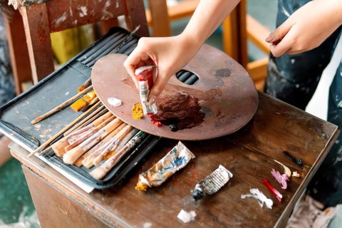 An artist's hand squeezes red paint onto a wooden palette, surrounded by brushes and used paint