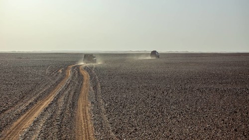 Two vehicles travelling on a dusty sandy road through a vast, open desert landscape, Off-road