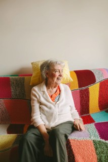 Elderly woman sits on a colorful, textured sofa, smiling warmly. She embodies warmth and joy,