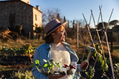 An woman in casual attire and a hat enjoys gardening under the sun, holding seedlings and a mini