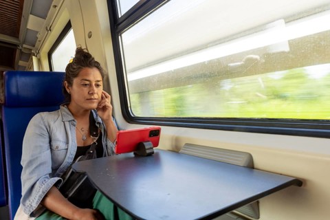 A woman sits by the window in a train, watching a video on her phone propped on the table The