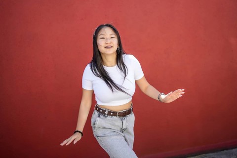 Cute urban female asian artist dancing K-pop dance smiling at camera outdoors against a red wall
