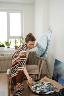 An artist in a cozy, light filled studio works on a canvas with paintbrushes and tubes nearby,
