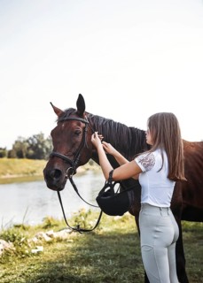 A young girl in equestrian attire prepares her horse by a peaceful river. The intimate connection