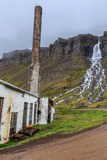 Old factory, fish factory, chimney, disused, waterfall and cliffs behind, cloudy, Djupavik,