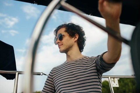 A man enjoys a peaceful afternoon sailing, steering a sailboat under clear skies. The striped shirt