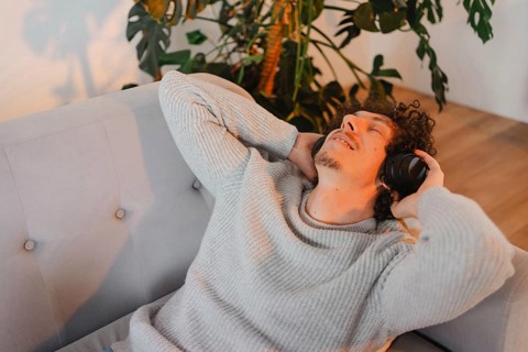A young man with curly hair relaxes on a sofa, listening to music with headphones. Sunlight streams