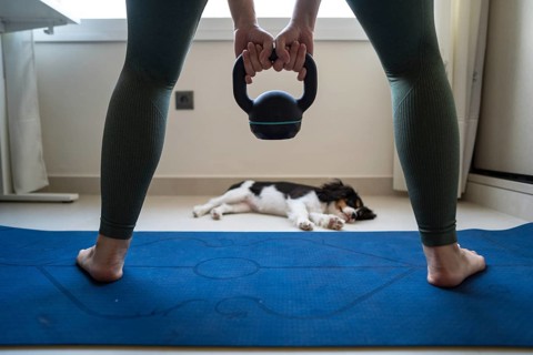 A young woman attempts to exercise at home, holding a kettlebell while her playful dog distracts