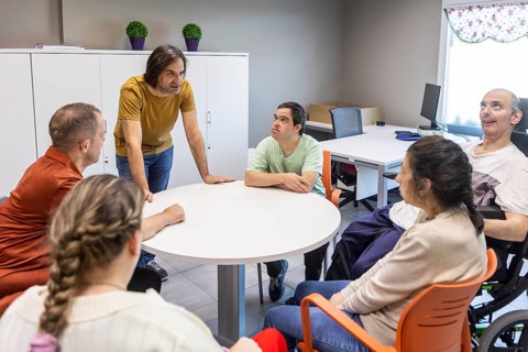 Individuals in a mental health day center engage in a group discussion around a table. The setting