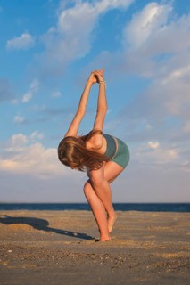 Woman practices standing Garudasana on beach at sunset, balancing with wrapped legs and crossed