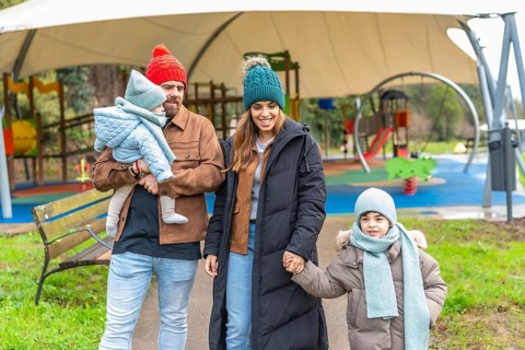 Smiling parents holding their children's hands are walking in a playground, wearing winter clothes