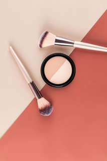 A pair of makeup brushes and a compact powder are artistically arranged on a split tone background