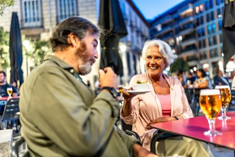 Senior man eating tapas next to partner in sidewalk cafeteria in the city of Madrid at night
