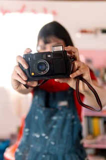 A young woman in a red shirt and paint-splattered overalls playfully holds a vintage camera, aiming