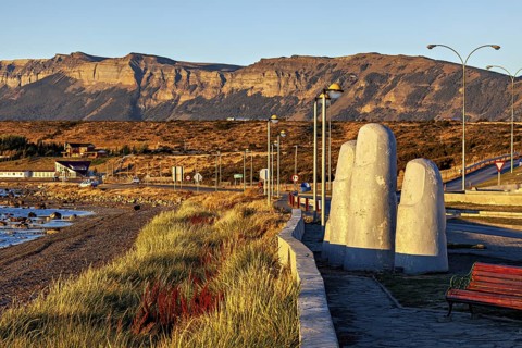A sculpture with large fingers next to a coastal road in a mountainous landscape in the evening