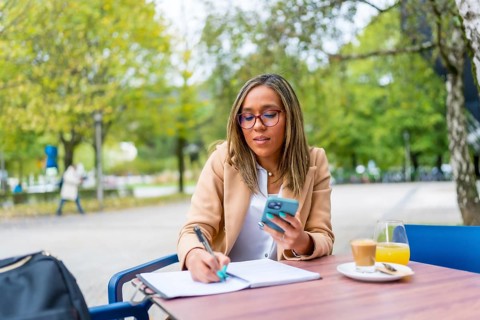 Latin businesswoman writing notes while working using mobile phone sitting in a sidewalk cafeteria
