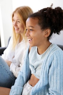 A joyful woman and girl share a laugh while sitting on a couch in a bright living room. The candid