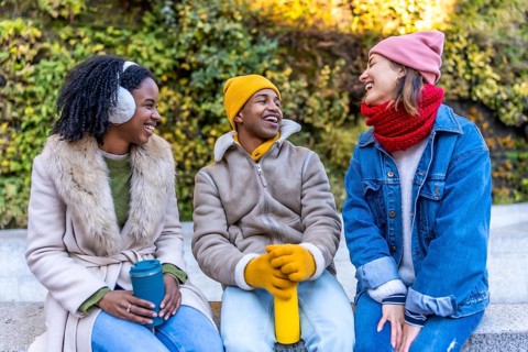 Three happy multi ethnic young friends are sitting and talking together in a city park during
