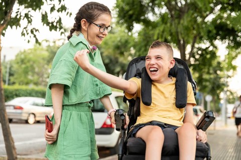A boy with cerebral palsy in a wheelchair and a girl with a tracheostomy laugh together outdoors,