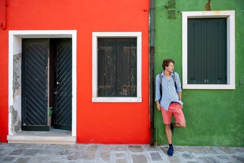 Young man with backpack leaning against a house wall, green and red house facade with entrance door