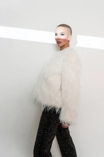 A confident middle-aged woman with a shaved head poses in a luxurious fur coat against a neutral
