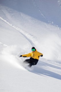 A snowboarder wearing a yellow jacket skillfully carves through fresh powder on a snowy mountain.