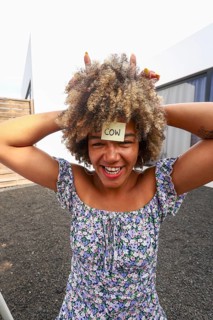 A joyful african american woman with a sticky note on her forehead labeled COW laughs while playing
