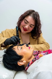 Beautician in black gloves and tan jacket smiles while performing precise permanent makeup
