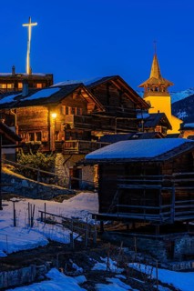 Snow-covered roofs of old farmhouses and barn storage, in the back the illuminated cross and the