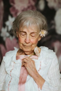 A peaceful portrait of an elderly woman in a white blouse, holding a flower, eyes gently closed.
