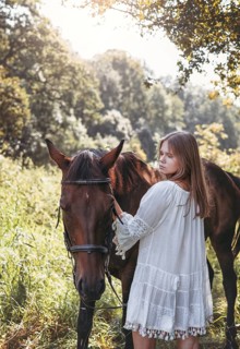 A young girl in a lace dress stands beside a horse in a lush, sunlit field. The scene captures the