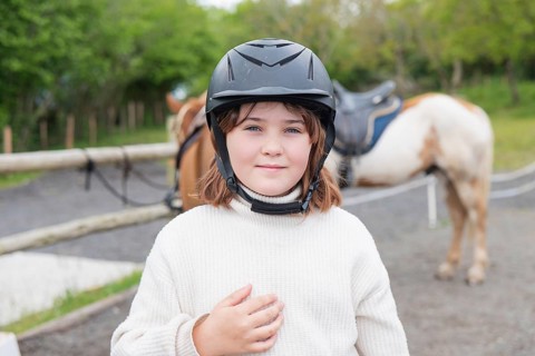 A young girl wearing a helmet enjoys a sunny day on the farm, standing beside her horse. The lush
