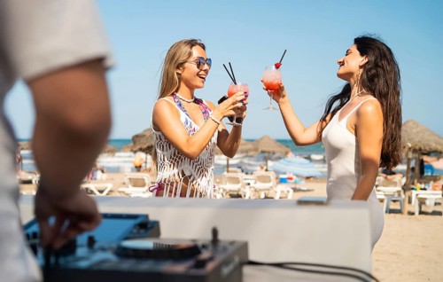 Two women enjoy colorful cocktails at a lively beach party. The sunny setting, energetic DJ music,
