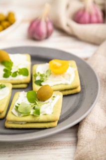 Green cracker sandwiches with cream cheese and cherry tomatoes on white wooden background and linen