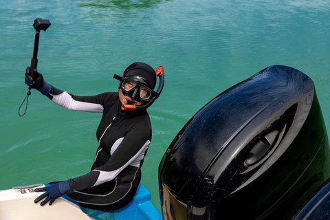 A snorkeler in a wetsuit and diving gear sits on a boat, holding an action camera, prepared for an