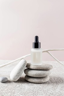 A minimalist skincare bottle with a black dropper cap rests on stacked stones against a neutral